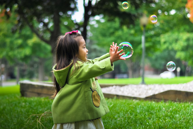 A girl in a green coat stands in a park trying to catch bubbles
