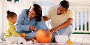 Two Black adults and a toddler prepare to carve a pumpkin on a front porch.